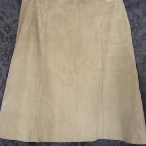 Könnte beinhalten: Ein beigefarbener Wildlederrock mit einer klaren A-Linien-Silhouette und vertikalen Nahtdetails auf der Vorderseite. Das Kleidungsstück hängt an einem Metallkleiderbügel mit zwei Clips und hat ein kleines weißes Markenetikett am Bund.