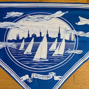 Könnte beinhalten: Blau-weißes Seidentuch mit nautischem Design. Das Tuch zeigt ein zentrales kreisförmiges Bild von Segelbooten auf dem Wasser mit einer Stadtkulisse im Hintergrund. Der Text "TALLINN '80" ist auf einem Banner zu sehen.