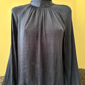 Può includere: Una camicetta a maniche lunghe blu scuro con collo alto. Il capo presenta uno scollo arricciato con pieghe verticali che scendono lungo il davanti, maniche lunghe con polsini e una finitura in tessuto liscio e leggermente riflettente.