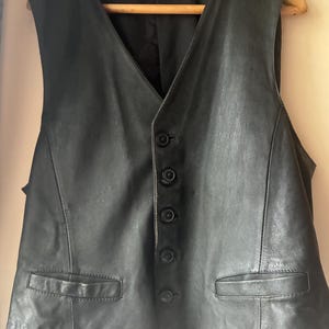 May include: A black leather vest featuring a V-neckline, a front button closure with five matching black buttons, and two horizontal welt pockets at the waist. The garment is sleeveless with a smooth finish and a structured, tailored appearance.
