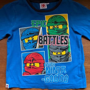 May include: A long-sleeved blue shirt featuring four ninja character portraits with different colored masks. The shirt has the words "EPIC BATTLES WITH NINJA" printed on it. The shirt has a black collar and a small Lego logo.