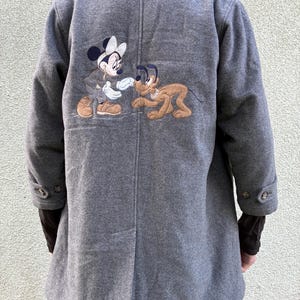 Könnte beinhalten: Ein grauer Mantel mit einer großen Stickerei auf der Rückseite, die die Zeichentrickfiguren Minnie Maus und Pluto darstellt. Der Mantel hat einen Kragen, lange Ärmel mit geknöpften Manschetten und eine vertikale Mittelnaht am Rücken.