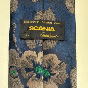 May include: A blue necktie featuring a floral pattern in shades of grey and brown. A black rectangular label is stitched to the center, displaying the text EXLUSIVE DESIGN FOR SCANIA BY Christian Jennes in yellow and white embroidery.