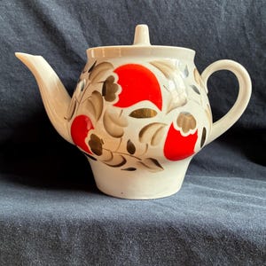 Può includere: Teiera in ceramica bianca con coperchio, caratterizzata da un motivo floreale con grandi fiori rossi, dettagli dorati e foglie marroni. Ha un beccuccio curvo e un manico arrotondato.