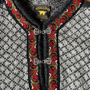 May include: A grey and white patterned knit sweater featuring a black trim with red and green floral embroidery along the neckline and front placket. The garment includes two silver metal clasps and a label reading VOLUND LOKKEN-NORWAY XL.