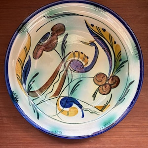 May include: A ceramic plate with a hand-painted design featuring birds and foliage. The plate has a blue rim and a light blue and white background. The birds are brown and purple, and the foliage is yellow, blue, and green.