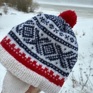 Puede incluir: Un gorro de invierno tejido con un patrón geométrico en azul marino y blanco, acentuado con rayas rojas y un pompón rojo. El gorro tiene orejeras y una cinta blanca. El gorro se muestra al aire libre en un entorno nevado.