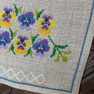 Peut inclure: Gros plan sur un tissu brodé au point de croix représentant des pensées colorées. Les fleurs sont dans les tons de jaune, violet et bleu, avec des tiges et des feuilles vertes. Le tissu a un fond beige avec une bordure bleue et des coutures décoratives blanches.