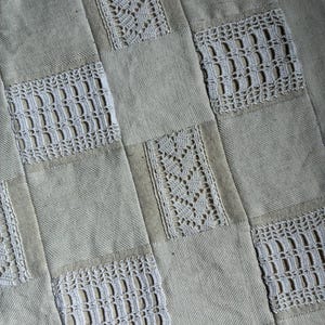 May include: A textile piece featuring a checkerboard pattern of alternating solid beige fabric squares and white crocheted lace panels. The lace panels display two distinct intricate openwork designs, creating a textured surface with a repeating grid layout.