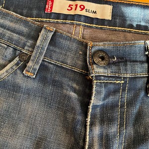 Puede incluir: Primer plano de unos vaqueros de mezclilla azules con cierre de botón y una etiqueta roja y blanca que dice "519 Slim". Los vaqueros tienen costuras amarillas y un diseño clásico de cinco bolsillos.