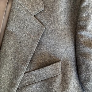 Pode incluir: Um blazer cinza-carvão com tecido texturizado semelhante a lã, com lapela entalhada, uma casa de botão na lapela esquerda e um bolso horizontal com aba na parte frontal inferior, complementado por um forro interno liso em cinza escuro.