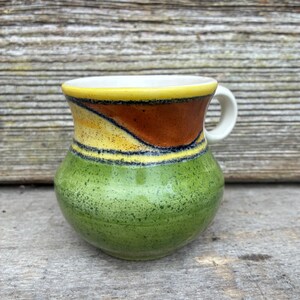 May include: A ceramic mug with a rounded body and a small handle. The upper portion features a yellow rim and a brown band with dark speckles. The lower portion is a vibrant green with a speckled texture throughout the glazed surface.