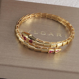 Puede incluir: Una pulsera dorada con forma de serpiente y diseño segmentado flexible. La cabeza y la cola están decoradas con pequeñas piedras preciosas rojas redondas. La pieza se muestra sobre una superficie marrón con el texto BVLGARI grabado.