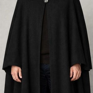 Puede incluir: Una capa de lana negra con cierre de botón en el cuello. La capa tiene un diseño amplio y fluido y se usa sobre un suéter oscuro y jeans azules. El modelo lleva una camisa con cuello debajo.