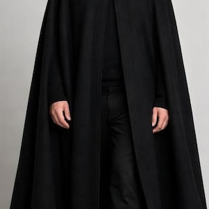May include: A long, black cape with a high collar and two buttons. The cape is worn over a black turtleneck and black pants. The person is wearing black shoes. The overall look is formal and elegant.