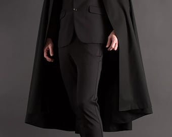 Puede incluir: Un traje negro con una capa a juego. El traje incluye una americana, pantalones de vestir y una corbata. La capa es larga y cubre los hombros y los brazos. El atuendo se completa con zapatos de vestir negros.