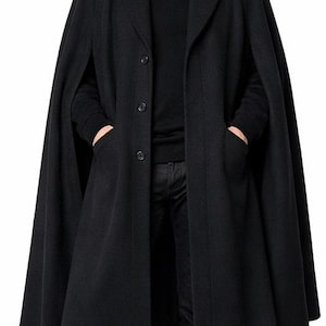 Op de afbeelding: Een zwarte wollen capejas met een knoopsluiting en een brede kraag. De jas heeft een lang, vloeiend ontwerp en wordt gedragen over een zwarte coltrui en een zwarte broek. Het model draagt zwarte schoenen.