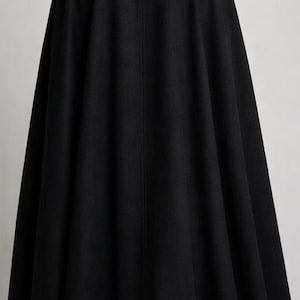 May include: A long, black cape with a high collar. The cape is made of a heavy fabric and falls to the ankles. The person is wearing black pants and black shoes. The cape has a vertical seam down the back.