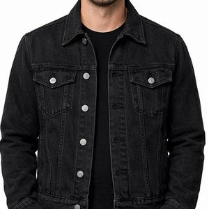 Puede incluir: Chaqueta vaquera negra con cuello clásico, cierre de botones y dos bolsillos en el pecho. La chaqueta tiene botones plateados y se lleva sobre una camiseta negra. El modelo tiene las manos en los bolsillos.