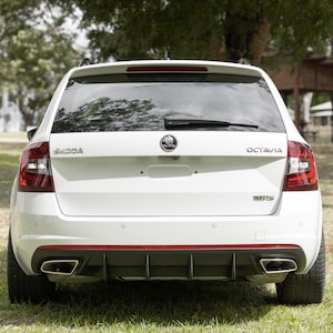 Könnte beinhalten: Rückansicht eines weißen Skoda Octavia Kombi. Das Auto hat schwarze Reifen, zwei Auspuffrohre und einen roten Akzent an der Stoßstange. Es steht auf einer Wiese mit Bäumen im Hintergrund.