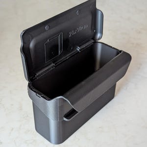 Puede incluir: Un contenedor de almacenamiento de plástico negro con una tapa abierta. La tapa está articulada y tiene las dimensiones "20x30cm" impresas. El contenedor es rectangular y se encuentra sobre una superficie clara.