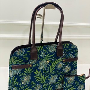 Può includere: Una borsa tote rettangolare e una piccola pochette abbinata con base in tessuto blu scuro e un motivo floreale ripetuto in verde e azzurro. La borsa presenta manici, finiture e una cinghia orizzontale marrone scuro con dettagli dorati.