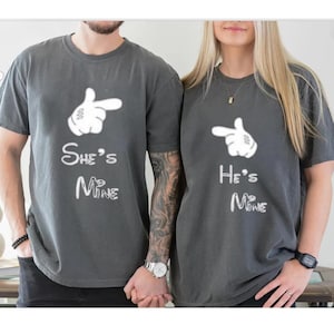May include: Two gray t-shirts with white graphic designs. One shirt says "She's Mine" and the other says "He's Mine". Each shirt features a white hand pointing to the side. The shirts are short-sleeved.