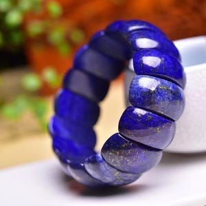 May include: A striking blue bracelet crafted from polished stones. The bracelet features a series of rounded, rectangular segments, each showcasing a deep, dark blue colour with delicate golden specks. The bracelet is presented on a white surface.