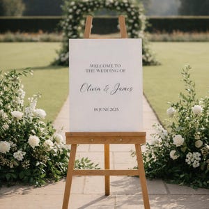 Könnte beinhalten: Eine Holzstaffelei trägt ein weißes Schild mit der Aufschrift "WELCOME TO THE WEDDING OF Olivia & James" und dem Datum "18. JUNI 2025". Das Schild steht auf einem Steinweg, umgeben von weißen Blumen und Grün, mit einem Blumenbogen im Hintergrund.