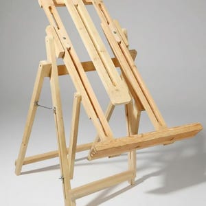 May include: A wooden artist's easel, ideal for painting or displaying artwork. The easel has an adjustable height and angle, with a robust base and a shelf for supplies. The light-coloured wood construction has a simple, functional design. The easel is made of natural wood.