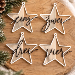 May include: Four star-shaped ornaments featuring a white outline, a light wood-toned centre with vertical slats, and black cursive text. The ornaments display the words eins, zwei, drei, and vier in a script font, each with a small circular loop at the top.