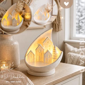 May include: A white, teardrop-shaped ceramic display featuring a miniature village scene with three small houses and a single pine tree. The interior is illuminated with a warm yellow light. The text 3D-Druckdatei STL / 3MF-Datei is visible.