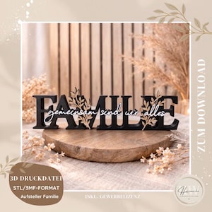以下が含まれることがあります： 「FAMILE」と書かれた黒い木製のサインで、「gemeinsam sind wir alles」という言葉が白い文字で書かれています。サインには装飾的な葉のアクセントがあり、木製の台座の上に置かれています。家族をテーマにした装飾品です。