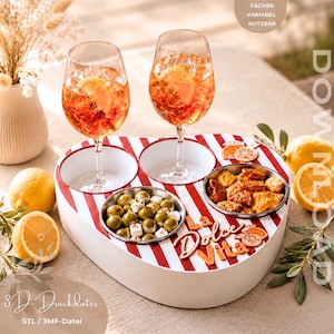 May include: A heart-shaped serving tray with red and white stripes, featuring two wine glasses filled with orange liquid, two metal bowls containing olives with cheese and crackers, and the text La Dolce Vita in orange script lettering.