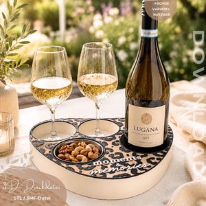 Könnte beinhalten: Ein herzförmiges Holztablett mit schwarzem Leopardenmuster, das zwei Weingläser, eine Flasche Lugana-Wein und eine kleine Metallschale mit Nüssen enthält. Auf dem Tablett steht der Schriftzug moments & memories in weißen Buchstaben.