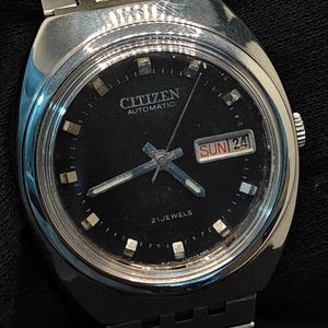 Pode incluir: Um relógio de pulso Citizen Automatic vintage com mostrador preto e ponteiros e marcadores prateados. O relógio apresenta um mostrador de dia e data e uma pulseira de aço inoxidável. O mostrador do relógio diz "Citizen Automatic" e "21 Jewels".