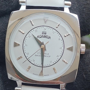 Könnte beinhalten: Eine Vintage-RW-Roamer-Uhr mit weißem Zifferblatt und silberfarbenem quadratischem Gehäuse. Die Uhr hat ein weißes Lederarmband. Das Zifferblatt zeigt den Text "RW ROAMER", "17 JEWELS SHOCKPROOF" und "SWISS MADE".