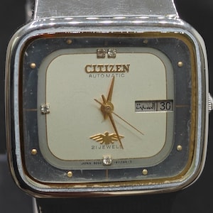 Puede incluir: Un reloj de pulsera automático Citizen vintage con caja cuadrada plateada y correa de cuero negro. La esfera crema tiene agujas e indicadores dorados y muestra la fecha. El reloj tiene el texto "Citizen Automatic" y "21 Jewels".