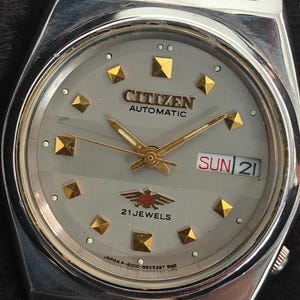 May include: A silver Citizen automatic wristwatch with a gray face. The watch features gold-colored hands and hour markers, and a day-date display. The watch face also displays the text "CITIZEN AUTOMATIC" and "21 JEWELS".