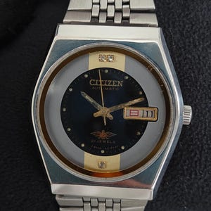 May include: A silver-tone Citizen automatic wristwatch with a dark blue circular dial, gold-tone accents, and two small clear stones at the top and bottom. The watch features a day-date window at the three o'clock position and a metal link band.