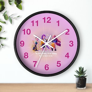 May include: A round wall clock with a black frame and a pink face. The clock face features a graphic of three figures and the text "No more hiding, I'll be shining, like I'm born to be." The numbers are in a darker pink.