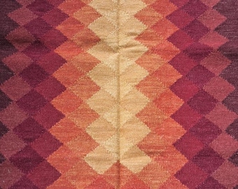Puede incluir: Una alfombra rectangular con un patrón de diamantes en tonos rojos, naranjas y beige. La alfombra tiene un borde con flecos y probablemente está hecha de material tejido.