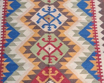 Puede incluir: Alfombra rectangular con un patrón geométrico en tonos rojos, azules, amarillos, verdes y beige. La alfombra tiene un borde con flecos y un diseño de diamantes repetido. La alfombra probablemente está hecha de tela tejida.