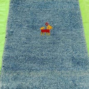 May include: A rectangular, blue bath mat with a textured surface. The mat features an embroidered design of a person on a horse in yellow, red, and orange. The mat is likely made of absorbent material.