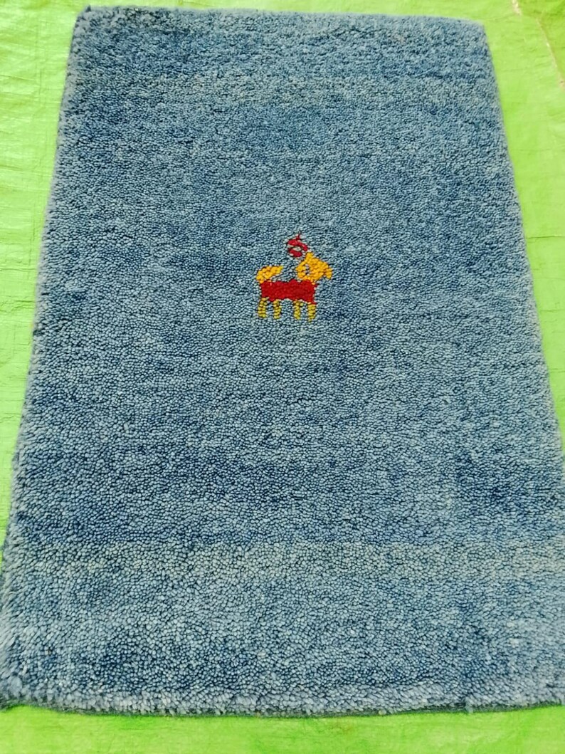 May include: A rectangular, blue bath mat with a textured surface. The mat features an embroidered design of a person on a horse in yellow, red, and orange. The mat is likely made of absorbent material.