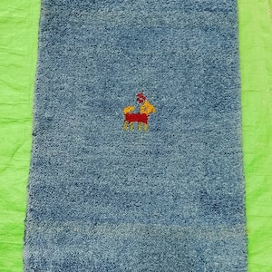 May include: A rectangular, blue bath mat with a small embroidered design of a horse in red, yellow, and white. The mat has a textured surface and is placed on a green surface.