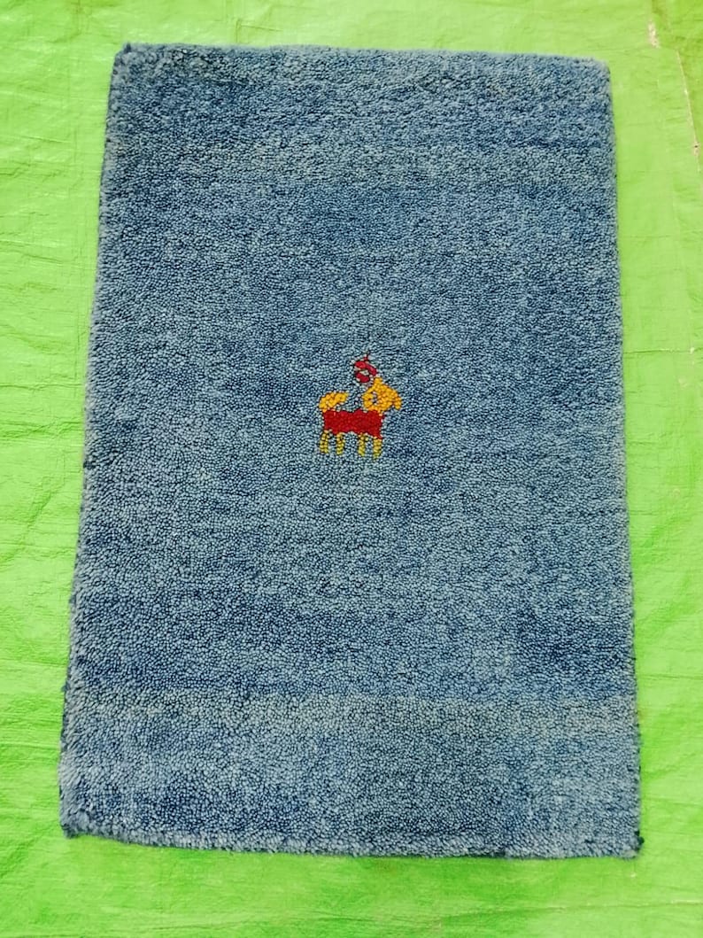 May include: A rectangular, blue bath mat with a small embroidered design of a horse in red, yellow, and white. The mat has a textured surface and is placed on a green surface.