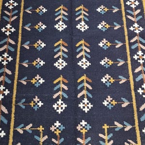 May include: A rectangular, dark blue rug with a repeating pattern of stylized floral and arrow motifs in white, tan, and blue. The rug has a tan border and fringed edges, suitable for home decor.