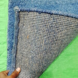 May include: A blue rectangular rug with a textured surface. The underside of the rug is a woven fabric in shades of blue and gray. The rug's edge is a darker blue, and the rug is on a green surface.