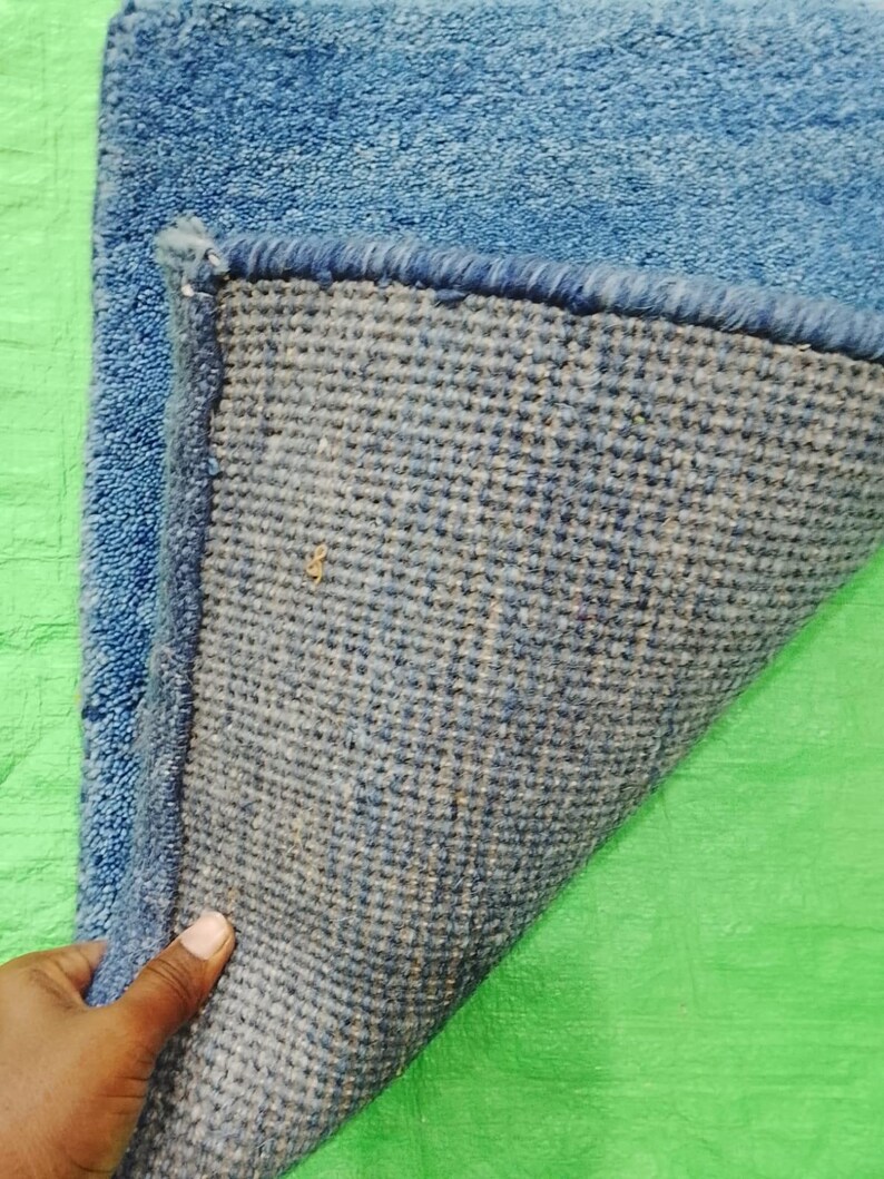 May include: A blue rectangular rug with a textured surface. The underside of the rug is a woven fabric in shades of blue and gray. The rug's edge is a darker blue, and the rug is on a green surface.
